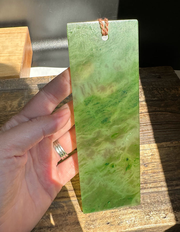 Six inch green jade bookmark being held by a hand, showing a marbled surface and a brown cord at the top.