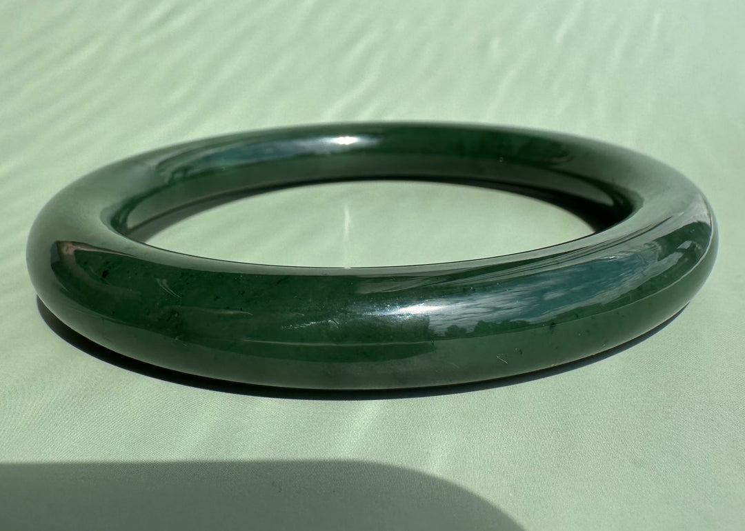 A dark green polished jade bangle bracelet resting on a light sage green textured surface.
