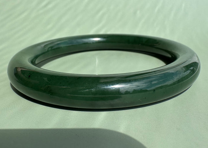 A dark green polished jade bangle bracelet resting on a light sage green textured surface.