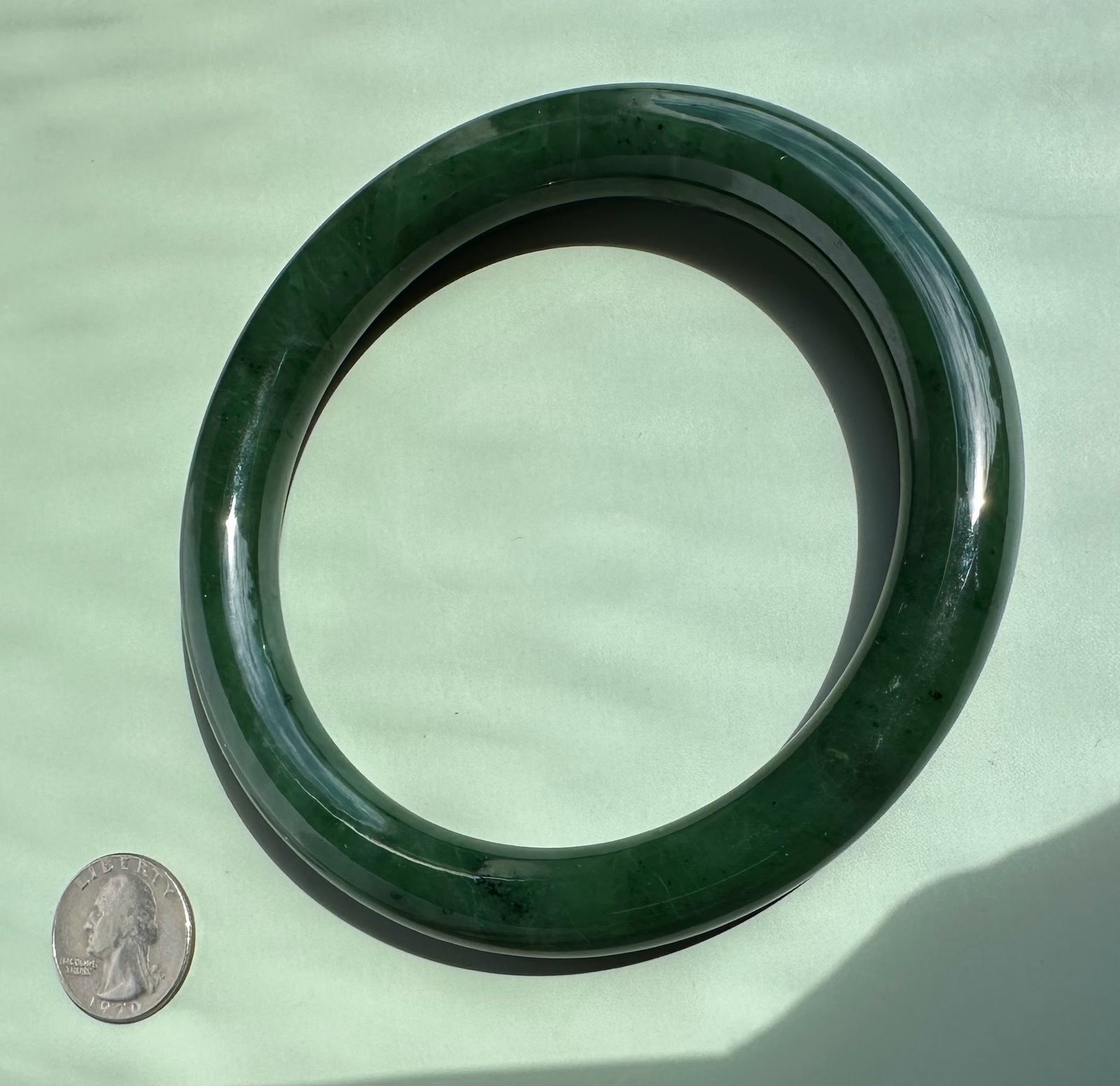 A round dark green jade bangle bracelet displayed on a light green surface, with a US quarter coin placed inside the ring to indicate scale.
