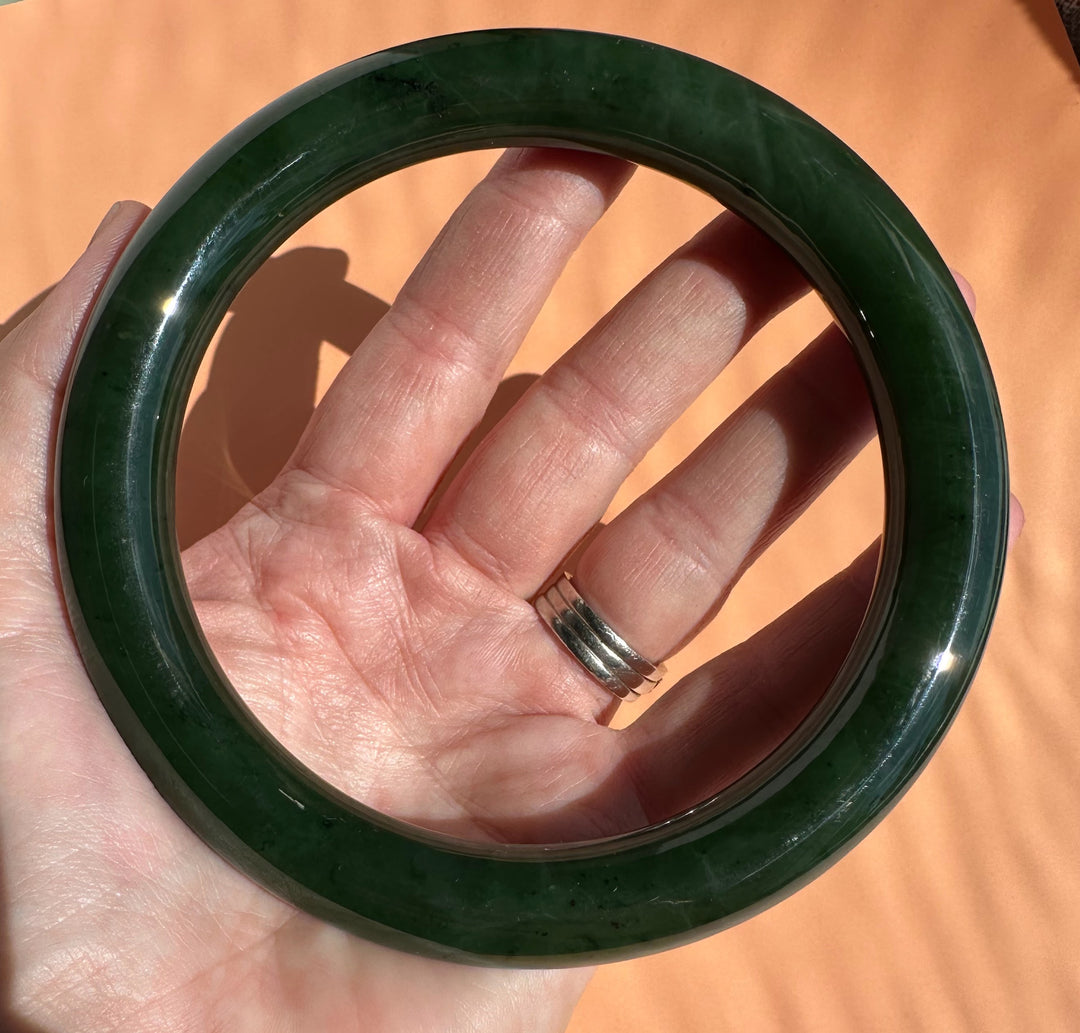 A dark green polished jade bangle held in a hand against a warm orange background.