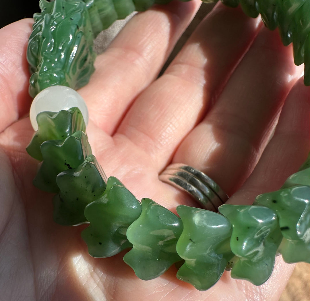 A close-up of a green carved beaded bracelet being held in a hand. The bracelet features carved jade-like green beads with a white round accent bead and a green dragon-shaped bead at one end.