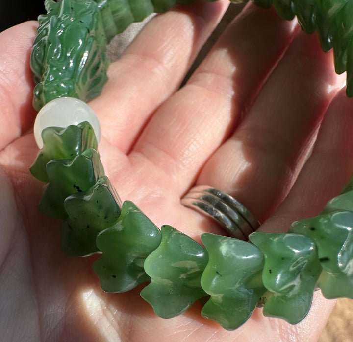A close-up of a green carved beaded bracelet being held in a hand. The bracelet features carved jade-like green beads with a white round accent bead and a green dragon-shaped bead at one end.