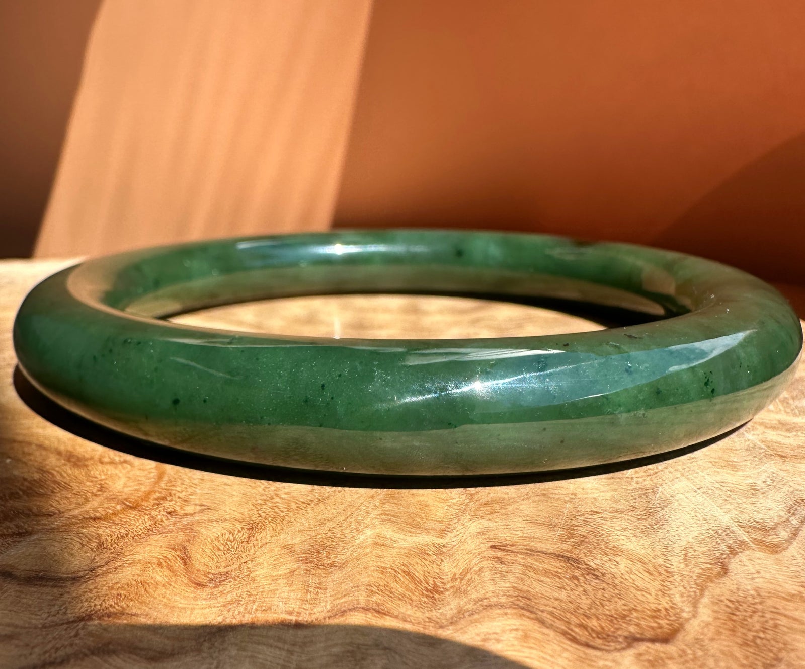 A polished green jade bangle bracelet resting on a textured wooden surface with a warm, orange-toned background.