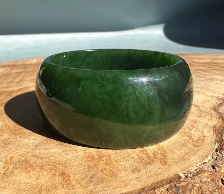 Wide Cassiar Jade Bangle in glossy deep green resting on a light wood surface.
