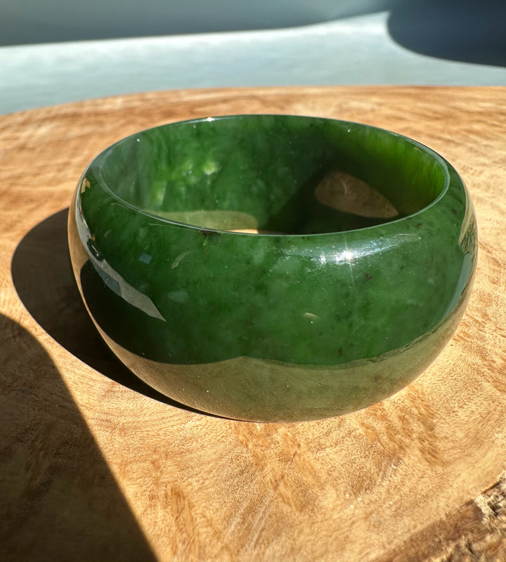 Wide Cassiar Jade Bangle with deep green color and glossy finish placed on a wooden surface