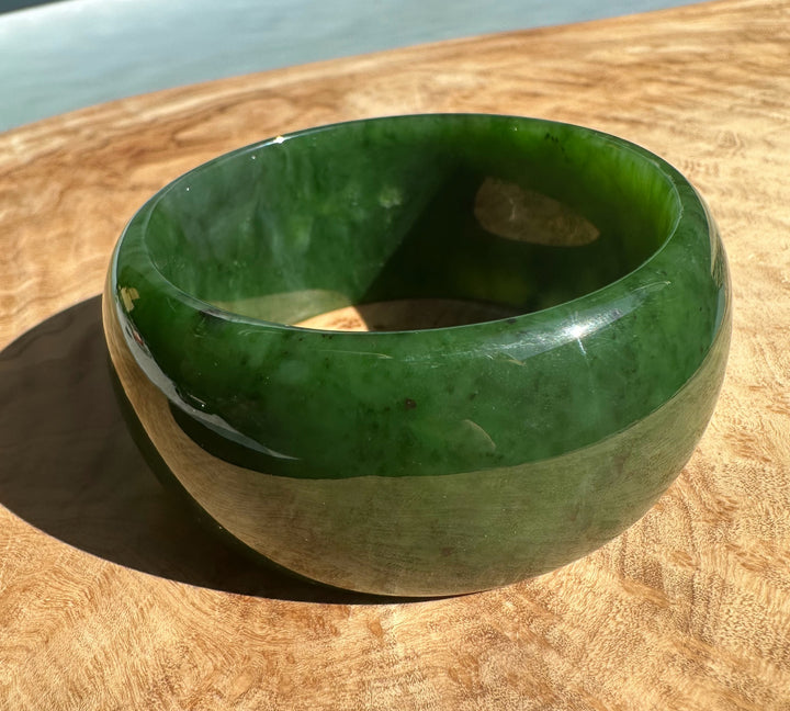 Side view of Wide Cassiar Jade Bangle resting on wood with glossy green jade and a beige lower band.