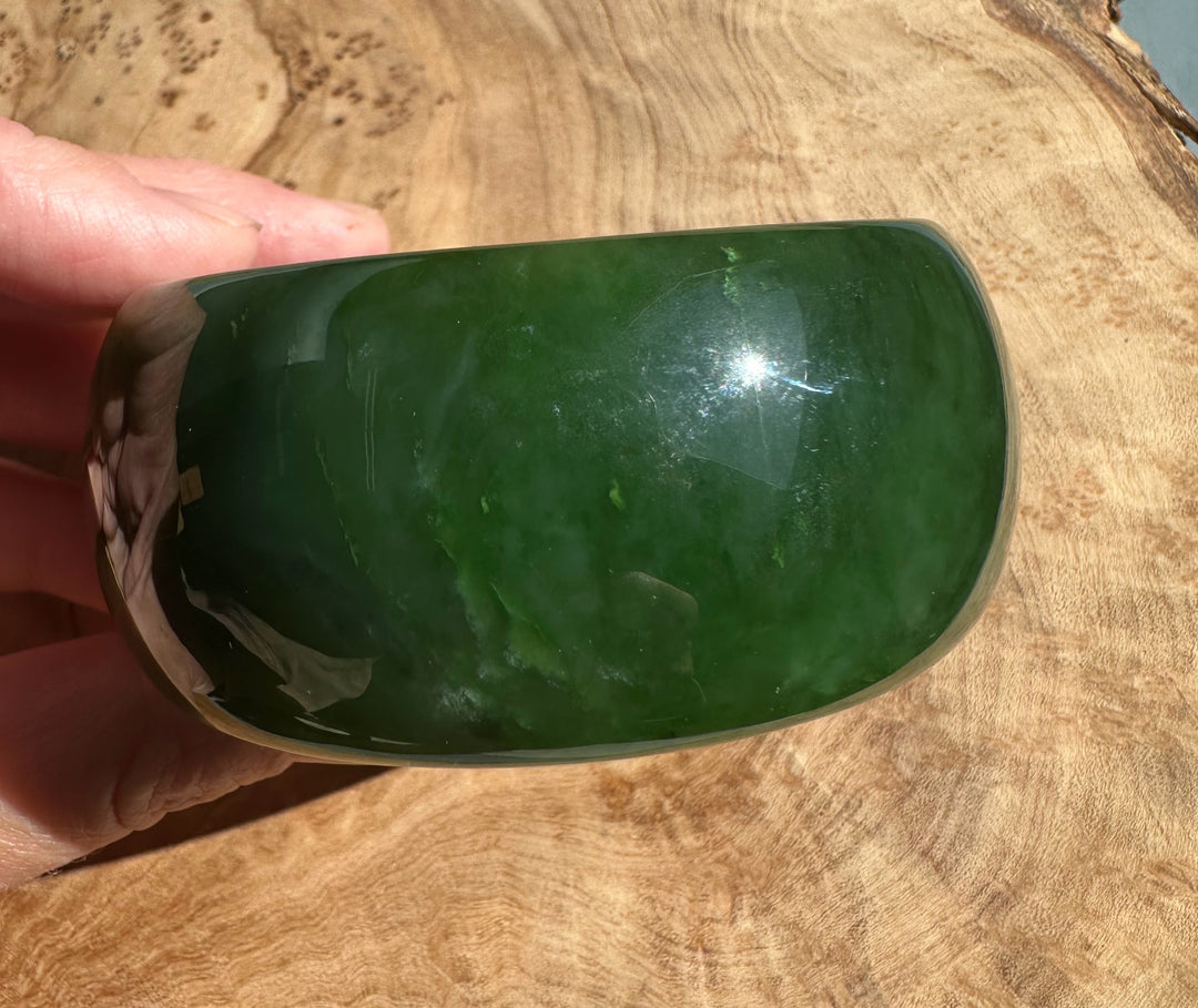 Wide Cassiar Jade Bangle 57.5 x 31mm in deep green with a polished surface held in fingers over a wooden surface