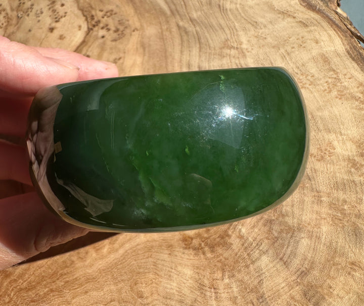 Wide Cassiar Jade Bangle 57.5 x 31mm in deep green with a polished surface held in fingers over a wooden surface