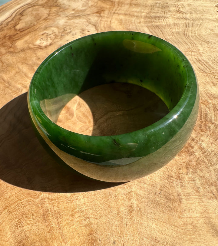 Wide Cassiar Jade Bangle 57.5 x 31mm resting on wood with glossy green exterior and visible inner edge