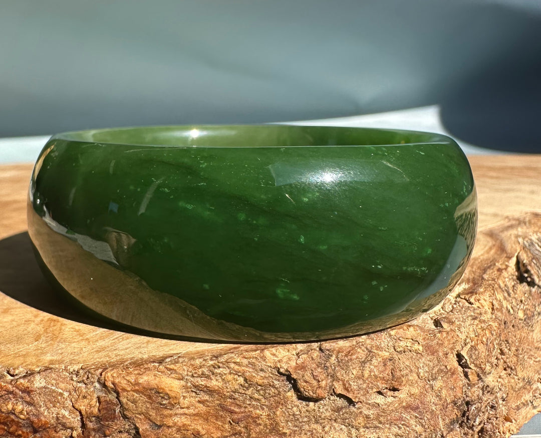 Wide Cassiar Jade Bangle in deep emerald green resting on a textured wooden surface.