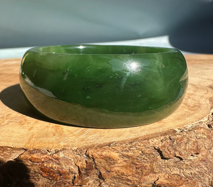 Wide Cassiar jade bangle with a smooth polished green finish resting on a wooden surface.
