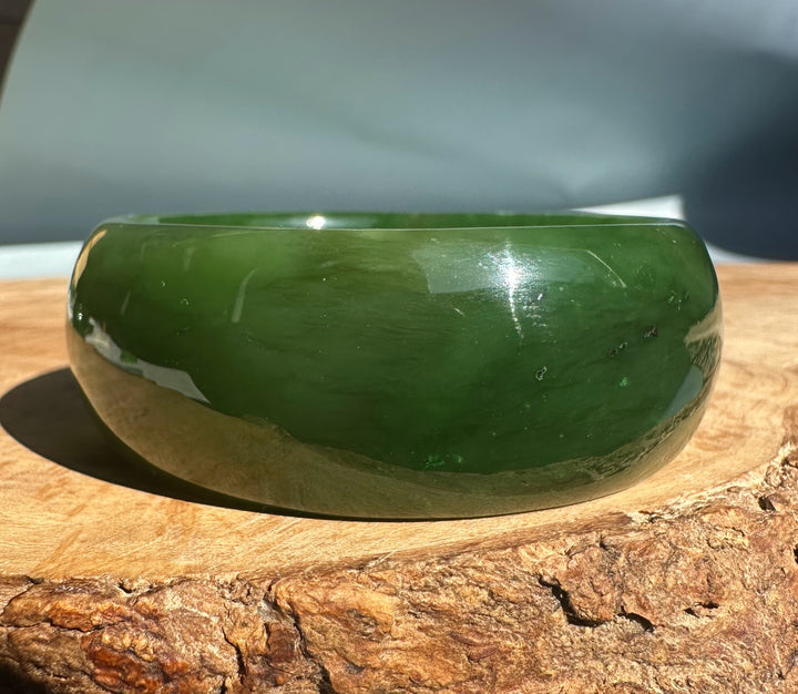 Wide Cassiar Jade Bangle in deep green with a glossy finish resting on a wooden surface