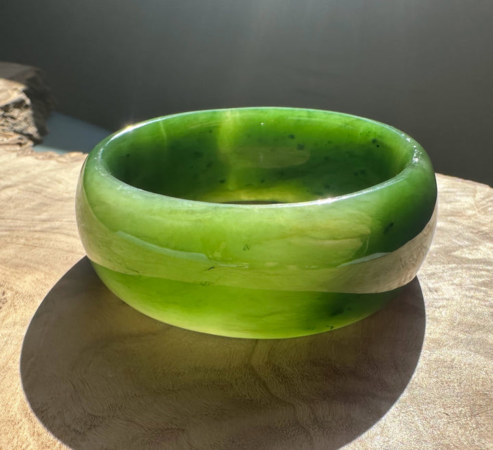 Wide Cassiar Jade Bangle 54 x 23.5mm in bright lime green with a pale band resting on a wooden surface