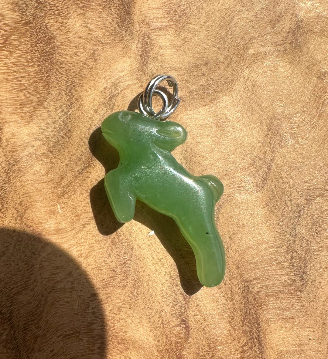 A green translucent jade pendant shaped like a rabbit, attached to a silver bail, resting on a textured brown wooden surface.