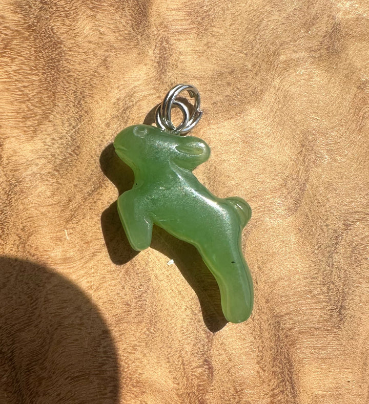 A green translucent jade pendant shaped like a rabbit, attached to a silver bail, resting on a textured brown wooden surface.