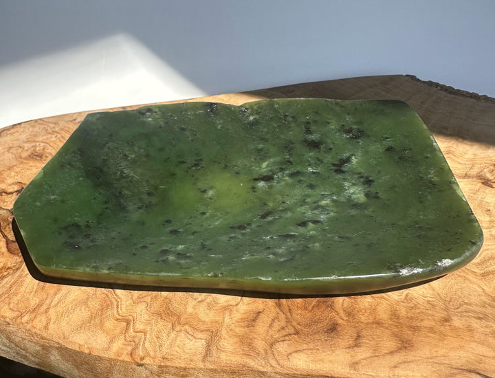 A polished green jade plate with natural dark inclusions, displayed on a rustic wood slice against a neutral background.