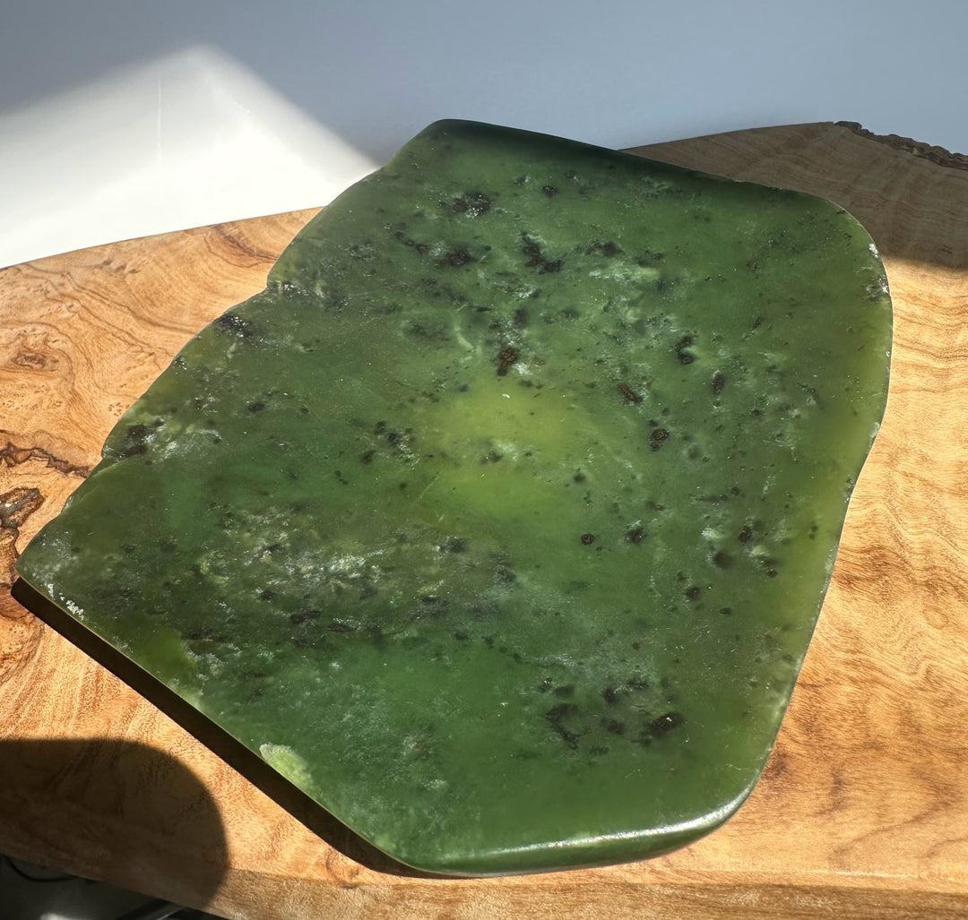 A polished green stone slab with natural dark inclusions, displayed on a wooden surface under direct sunlight.
