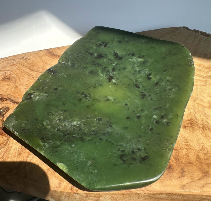 A polished green stone slab with natural dark inclusions, displayed on a wooden surface under direct sunlight.
