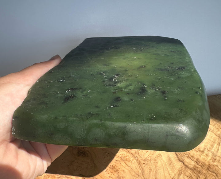A hand holding a thick, polished green stone slab with a rounded rectangular shape, showing natural marbling and dark inclusions, set against a neutral background.
