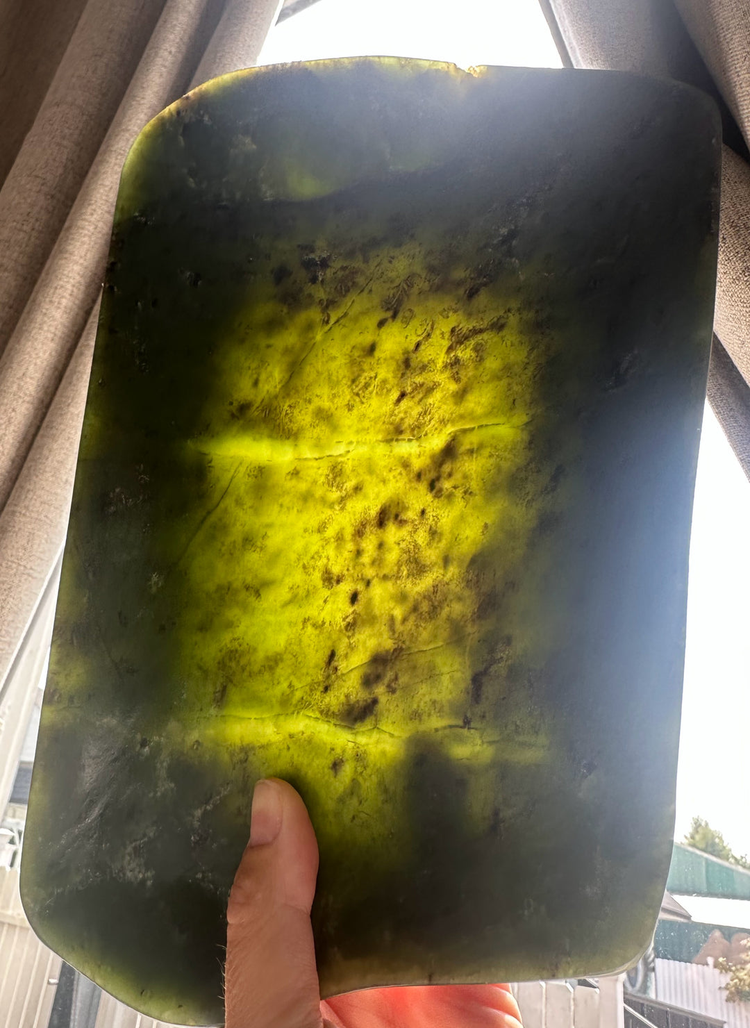 A hand holding a large, polished rectangular stone slab with rounded corners, showing a natural marbled pattern of bright yellow-green, dark green, and brownish inclusions, photographed in natural light near a window.