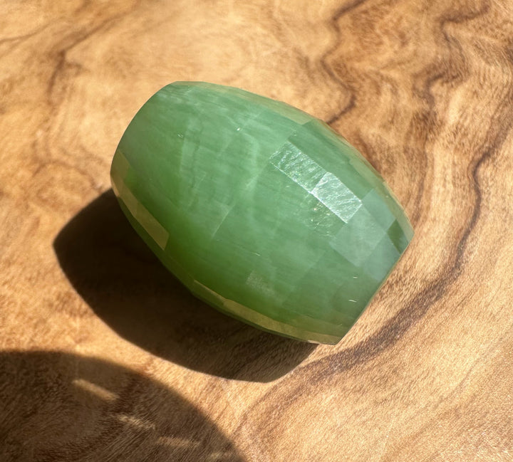 A faceted green jade gemstone with a rounded teardrop shape, resting on a textured wooden surface.