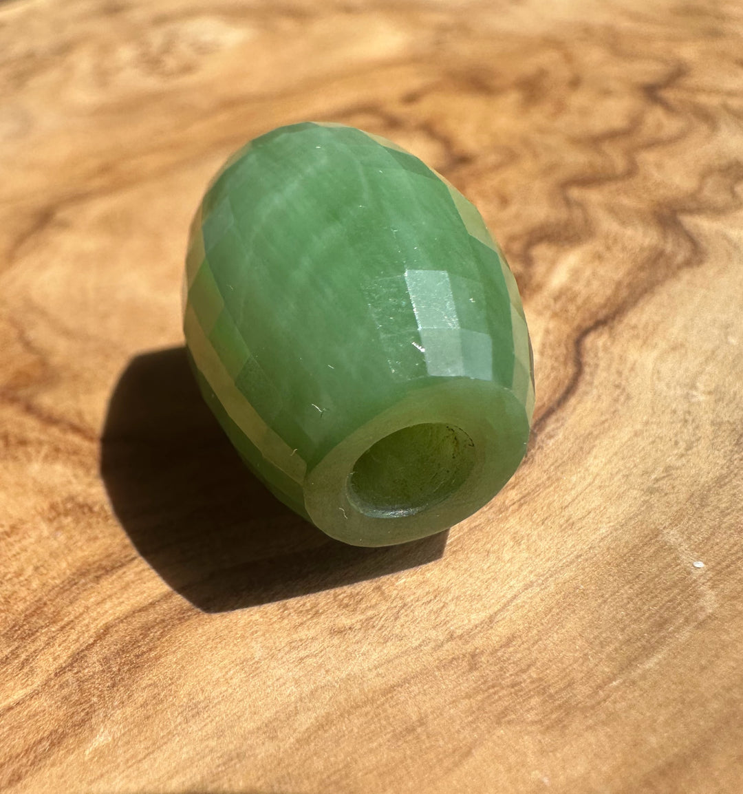 A faceted green stone bead with a central hole, resting on a textured wooden surface.