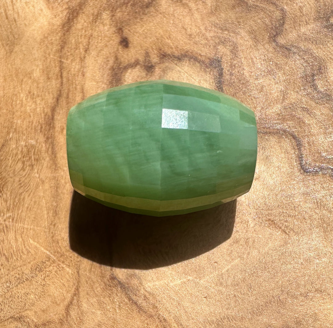 A faceted green stone bead, jade piece, resting on a textured wooden surface.