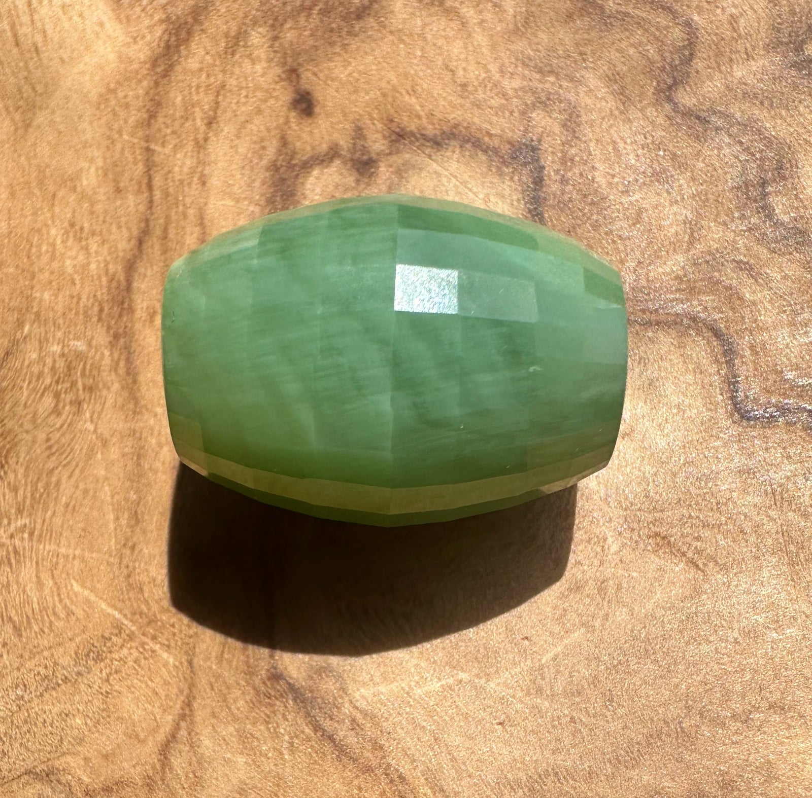 A faceted green stone bead, jade piece, resting on a textured wooden surface.