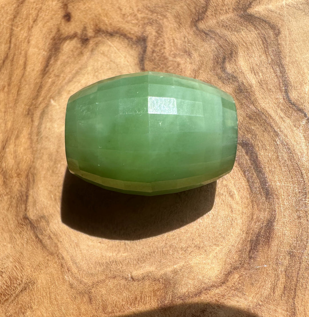 A faceted green stone bead,jade, resting on a textured wooden surface.