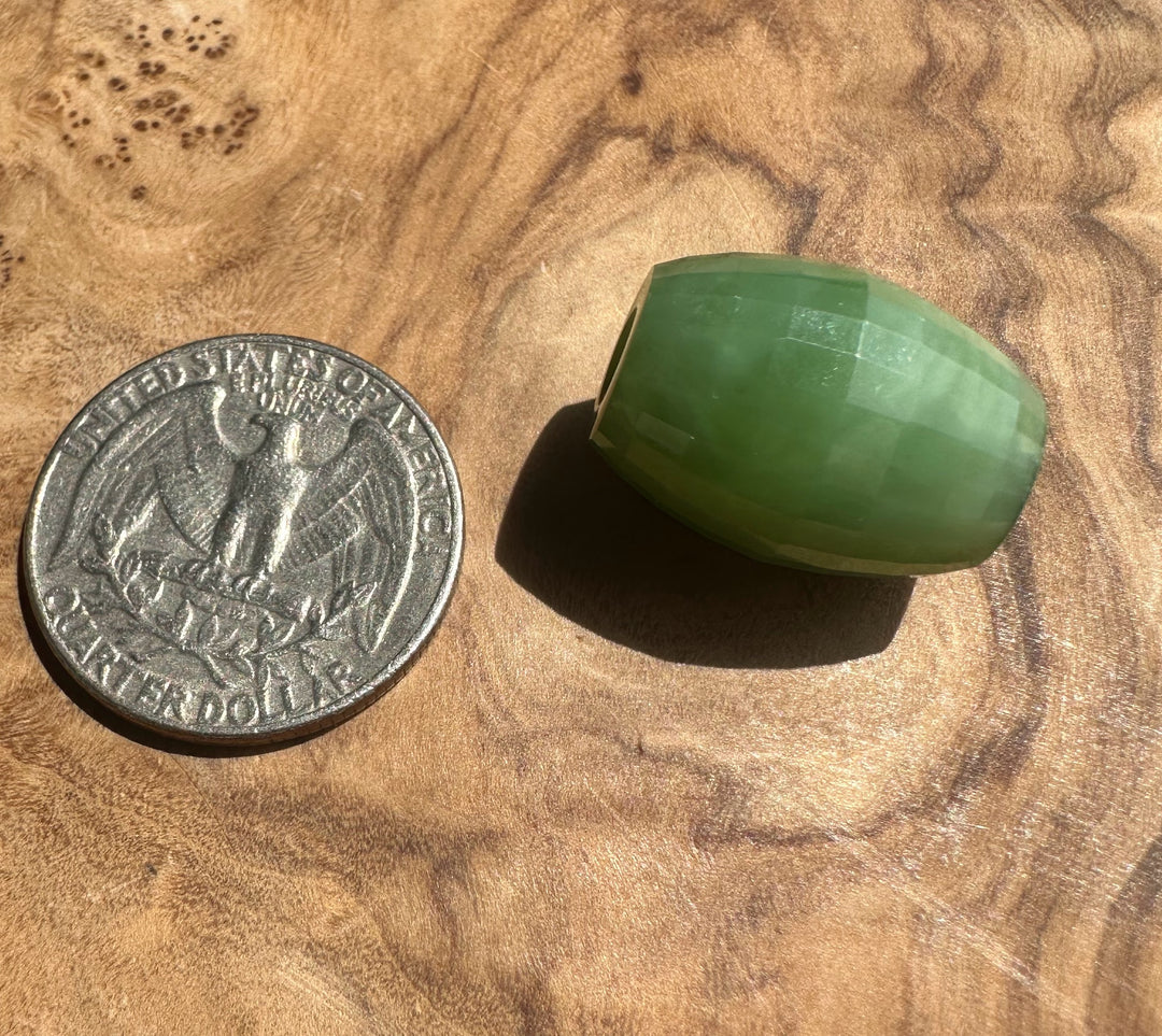 A faceted green stone bead,  jade, shown next to a US quarter coin for scale on a textured wooden surface.