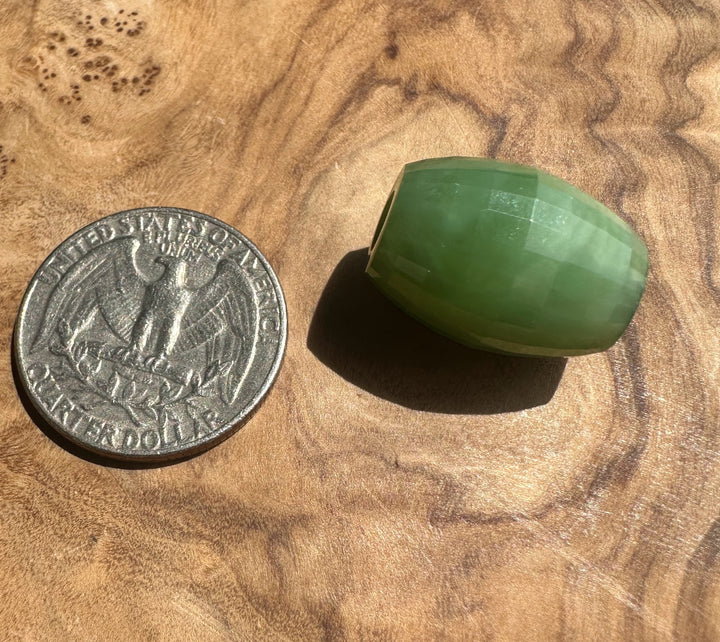 A faceted green stone bead,  jade, shown next to a US quarter coin for scale on a textured wooden surface.