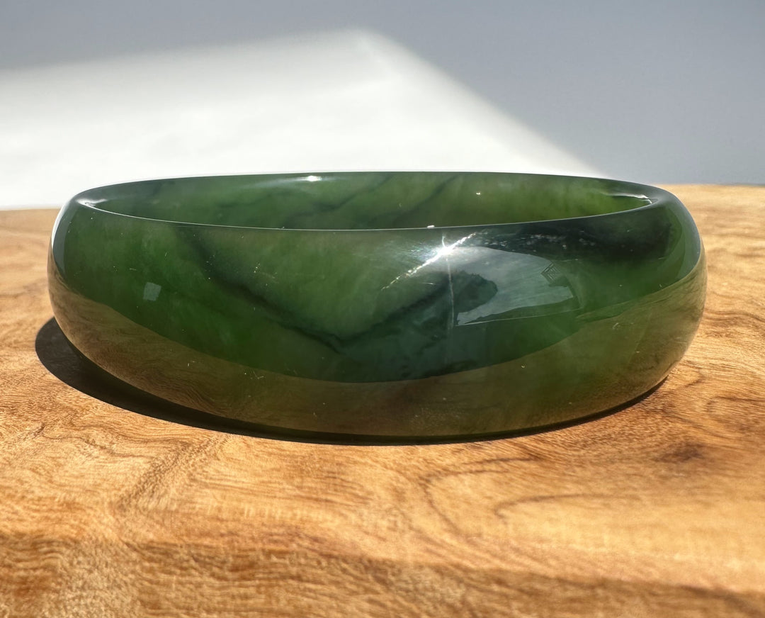 A polished dark green jade bangle bracelet resting on a wooden surface with a white background behind it.