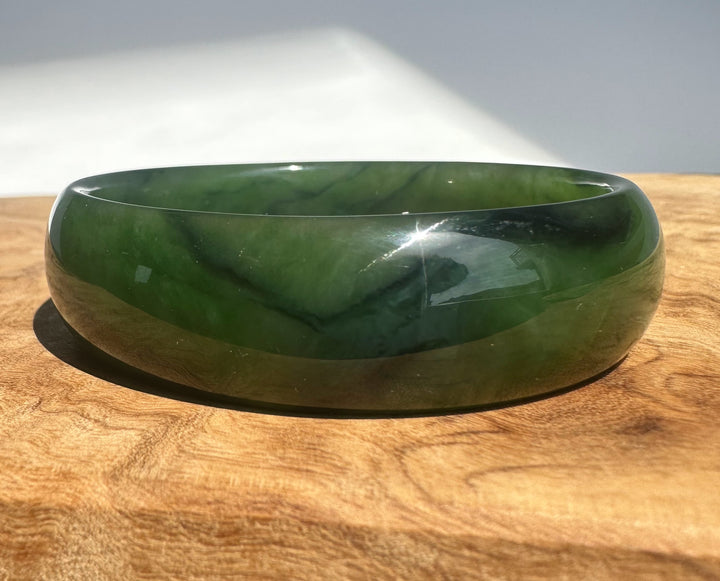 A polished dark green jade bangle bracelet resting on a wooden surface with a white background behind it.