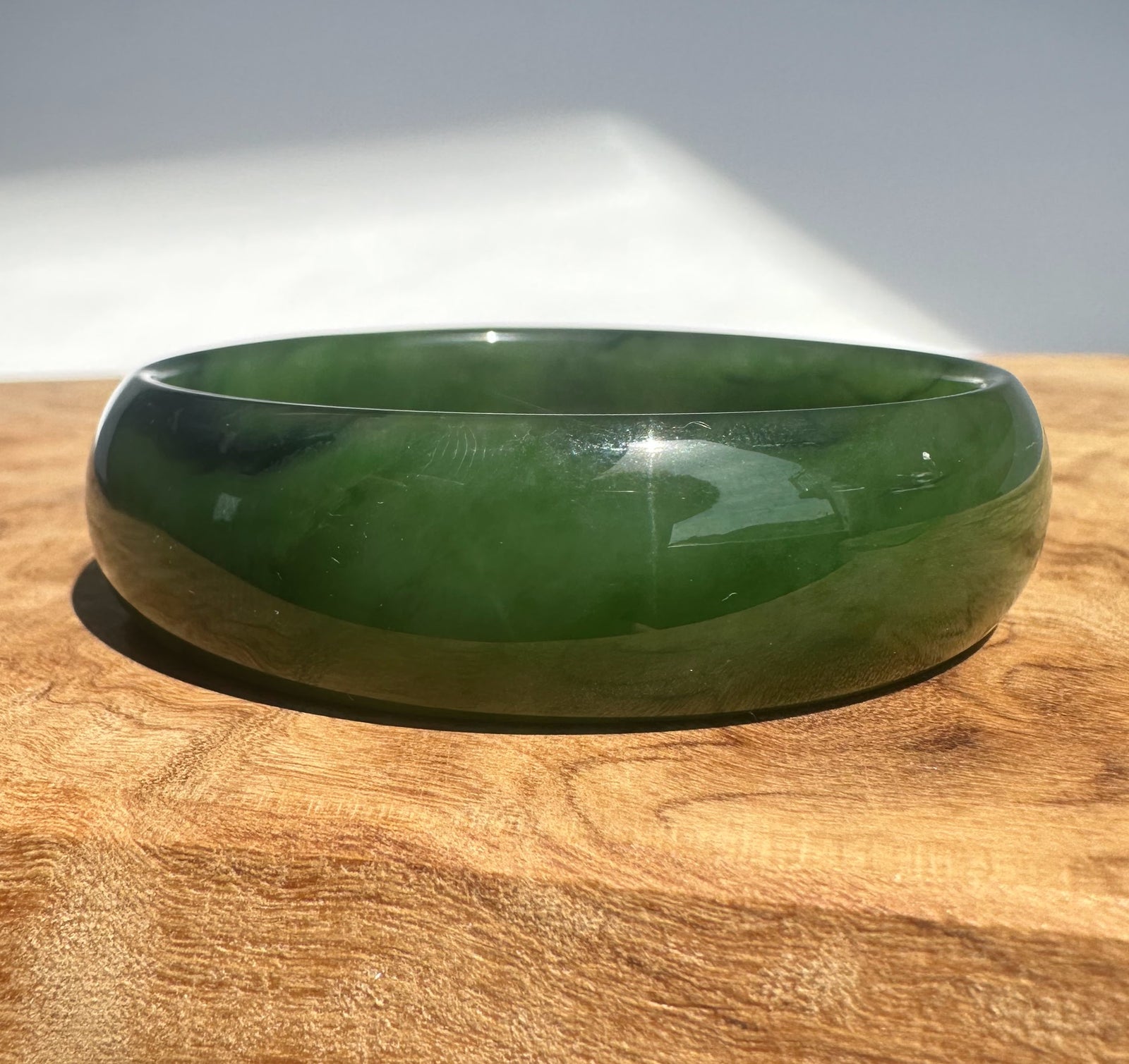 A polished dark green jade bangle bracelet resting on a wooden surface with a soft-focus gray background.