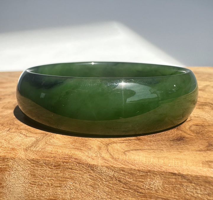 A polished dark green jade bangle bracelet resting on a wooden surface with a soft-focus gray background.