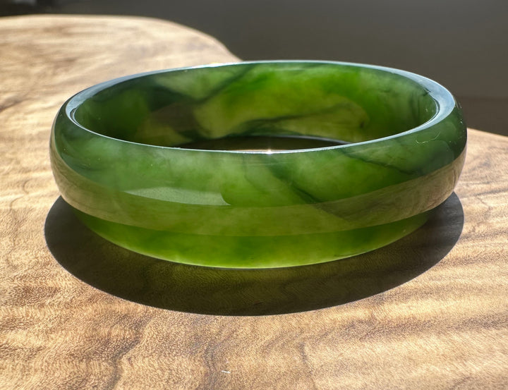 A polished green jade bangle bracelet with natural marbled patterns, resting on a textured wooden surface.