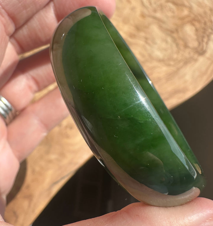 A hand holding a polished dark green jade bangle bracelet against a wooden background.
