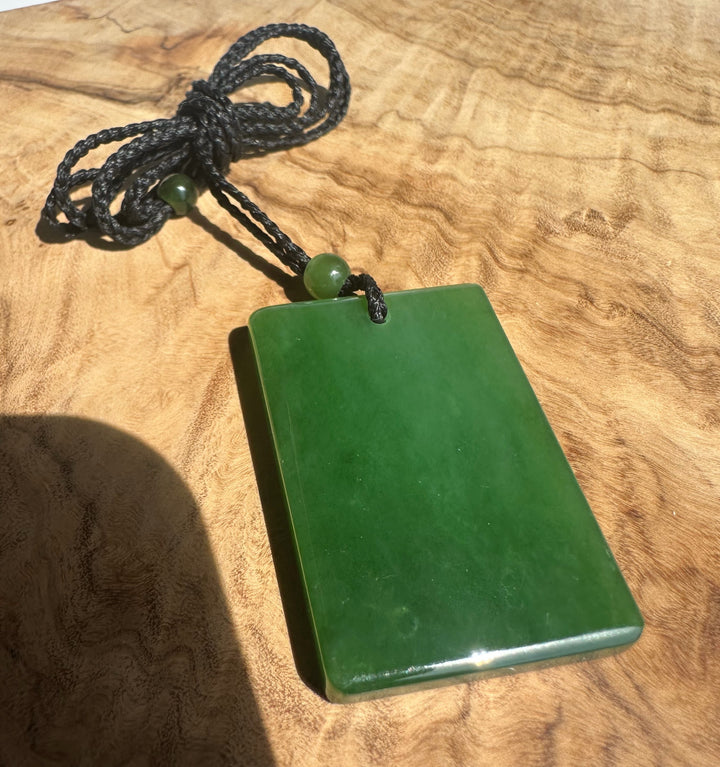 A green polished stone pendant with a dark braided cord and a small green bead, displayed on a wooden surface.