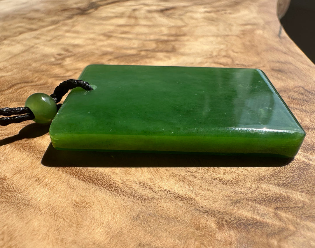 A green polished stone pendant or plaque with a black cord and round green bead, resting on a wooden surface.