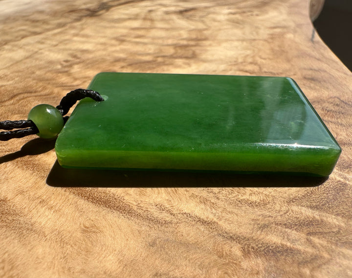 A green polished stone pendant or plaque with a black cord and round green bead, resting on a wooden surface.