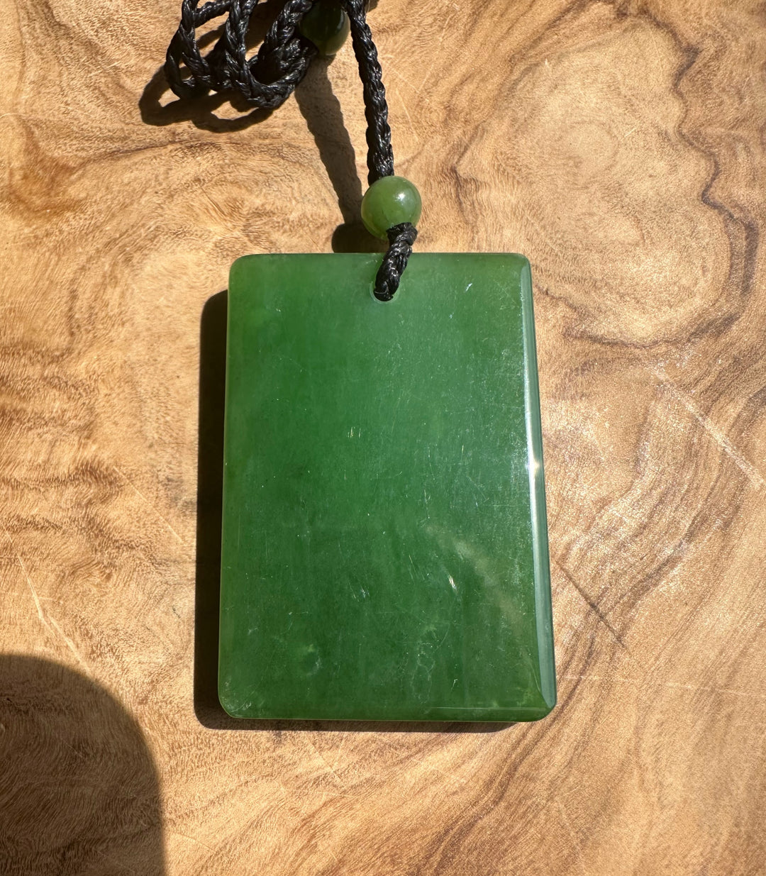 A polished green jade pendant necklace with a black braided cord and a small green bead, displayed on a textured wooden surface.