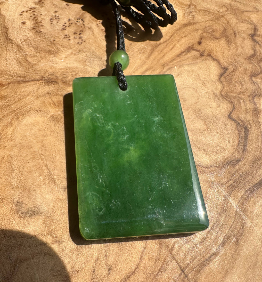 A polished green stone pendant necklace with a rectangular shape and a small green bead, resting on a textured wooden surface.