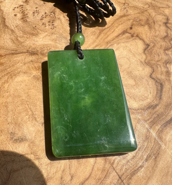 A polished green stone pendant necklace with a rectangular shape and a small green bead, resting on a textured wooden surface.