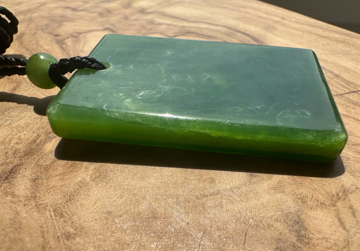 A rectangular green jade pendant with a black cord and a small green jade, resting on a wooden surface.