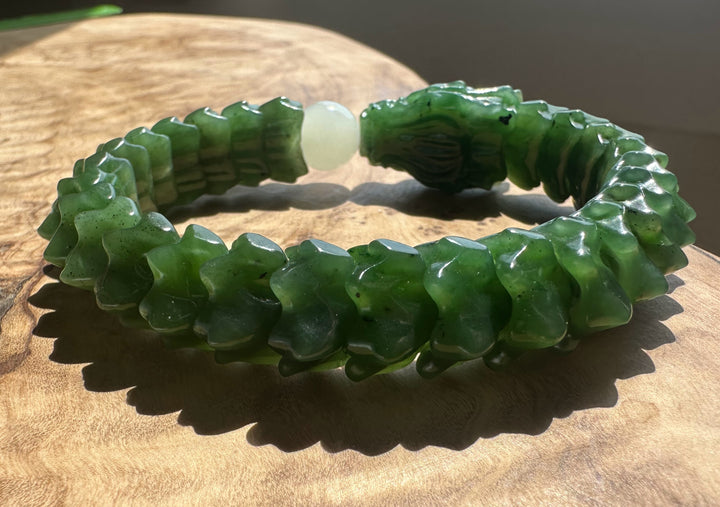 A carved green jade bracelet with a detailed dragon head closure and a central white bead, displayed on a wooden surface.