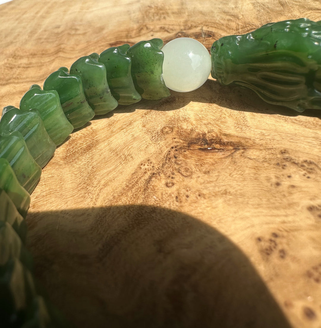 A close-up of a green jade-like beaded bracelet with carved dragon head ends and a round white bead, laid on a wooden surface.