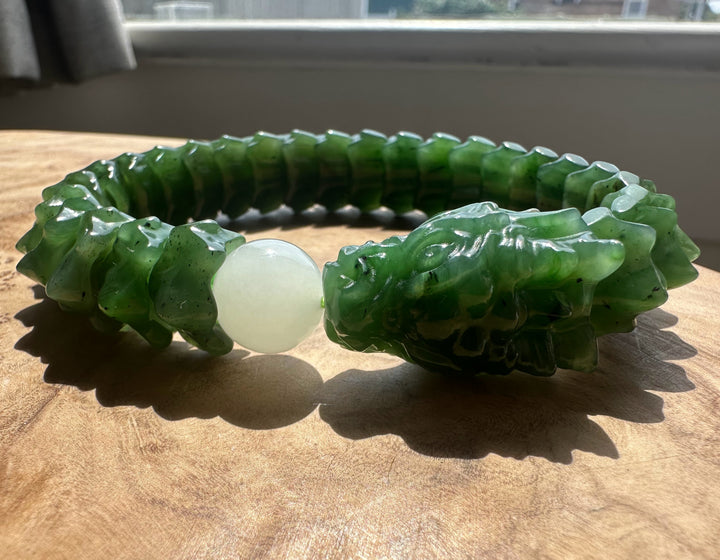 A carved green jade bracelet with a dragon head closure and a smooth white bead, displayed on a wooden surface near a window.
