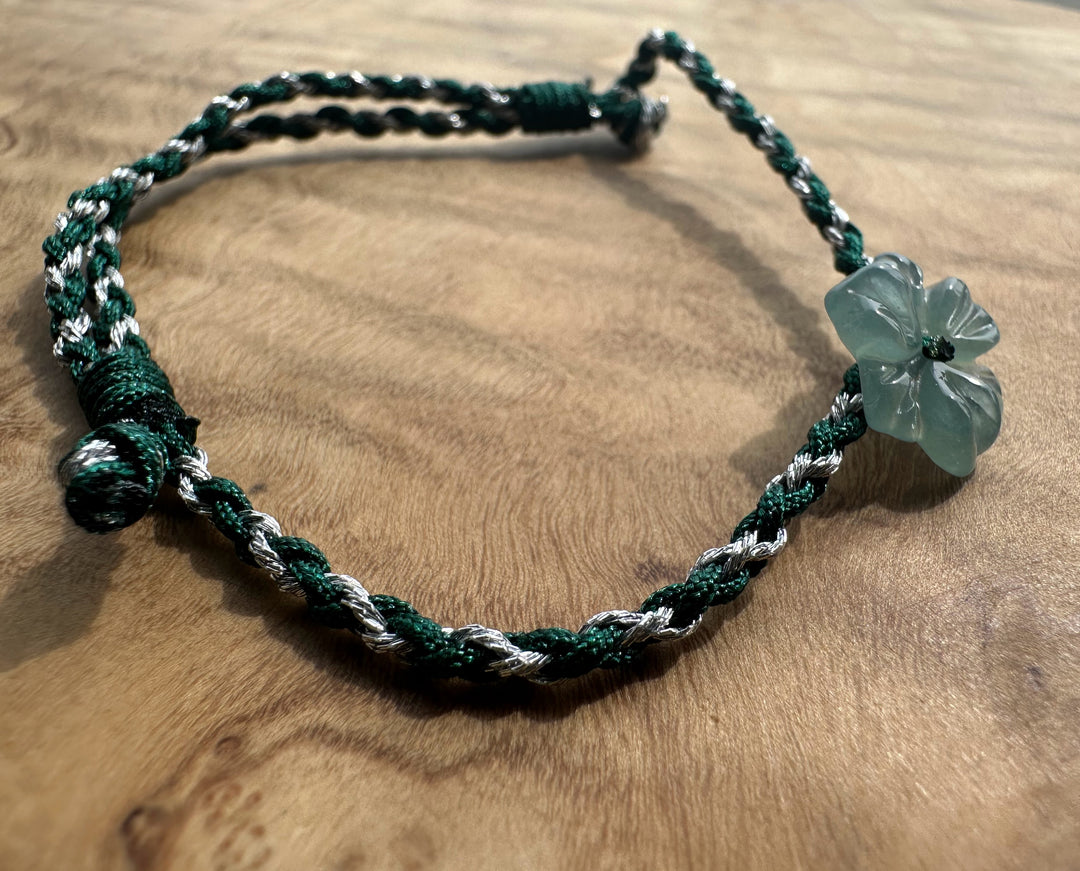 A handmade braided cord bracelet in dark green and white, featuring a translucent light green four-petal flower charm, displayed on a wooden surface.