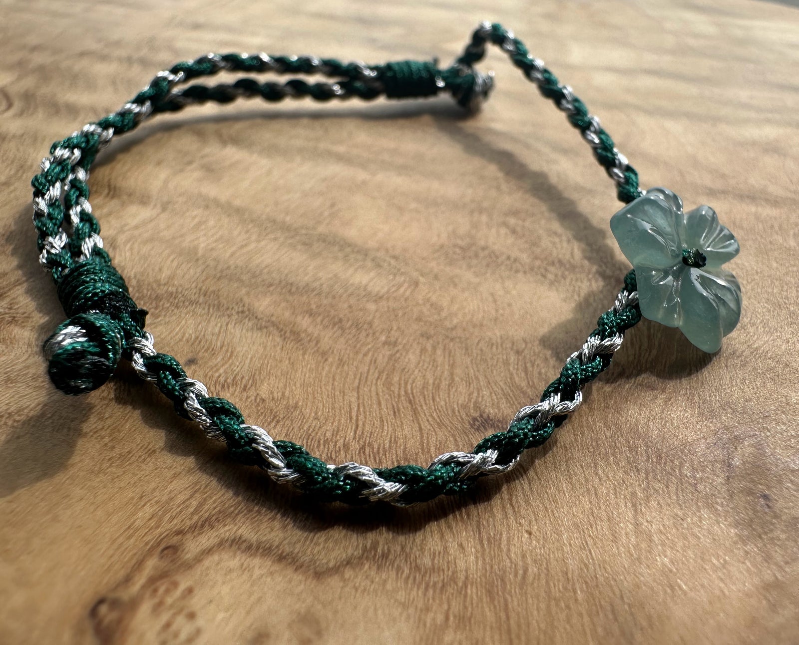 A handmade braided cord bracelet in dark green and white, featuring a translucent light green four-petal flower charm, displayed on a wooden surface.